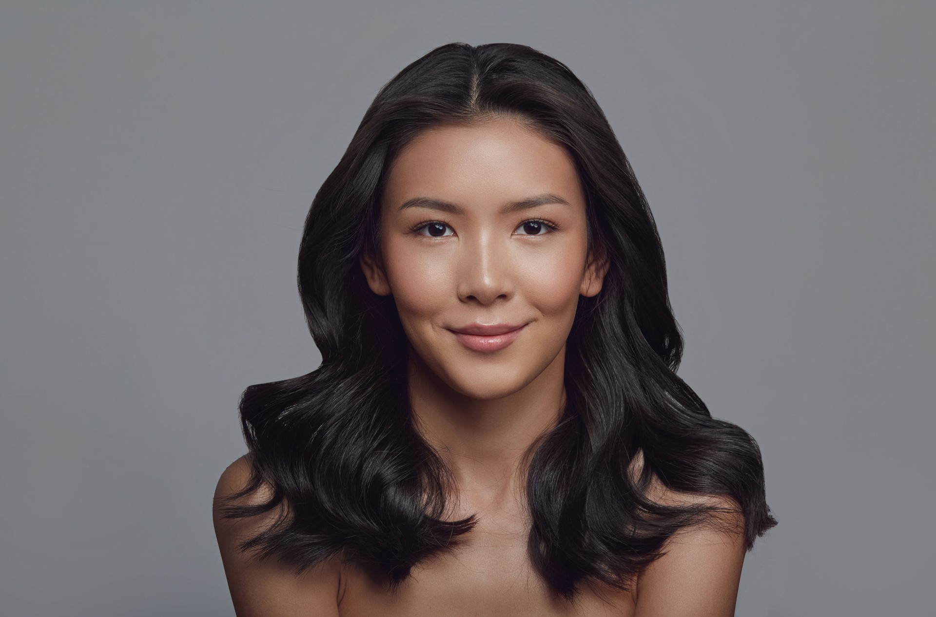 Beautiful young asian brunette woman with clean fresh skin and wavy healthy hair on gray background.